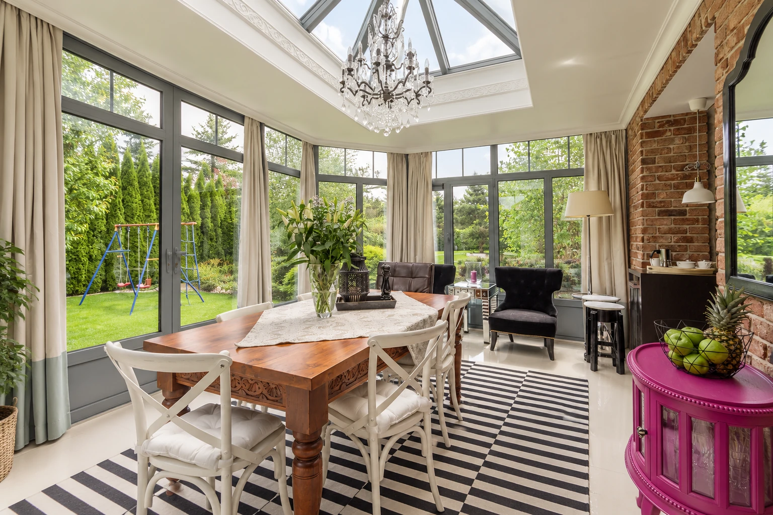 Bright all-season room with a wooden dining table, white chairs, skylight, chandelier, and large windows overlooking a garden.