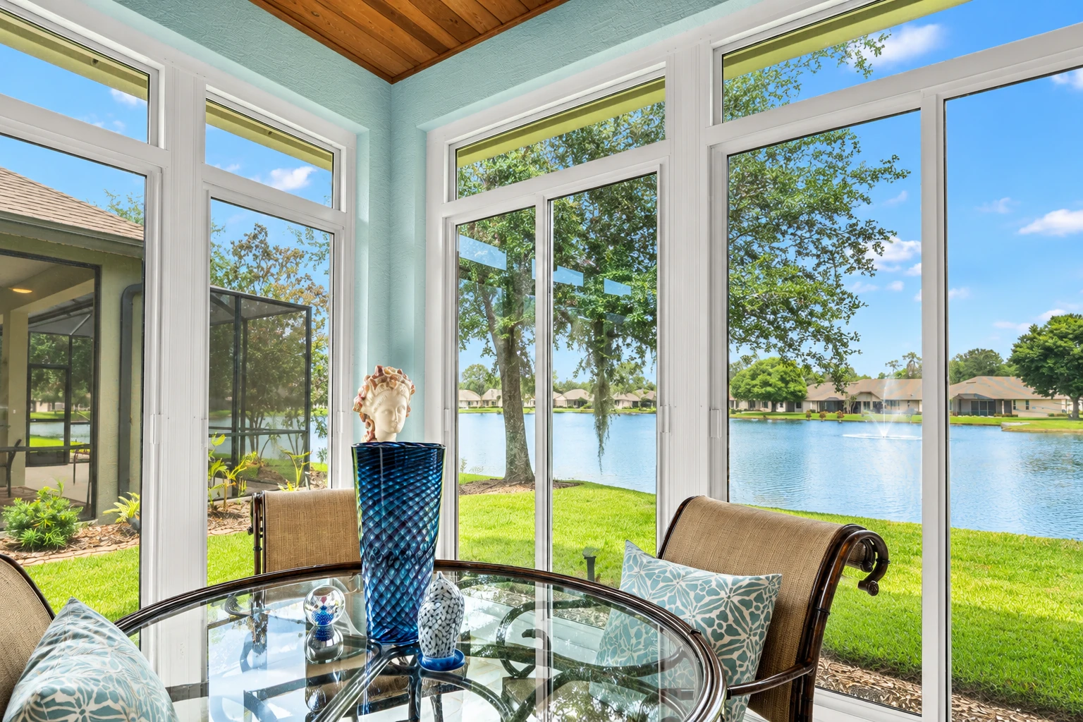 Custom sunroom with tall white-framed windows, a glass dining table and views of a lake and lawn.