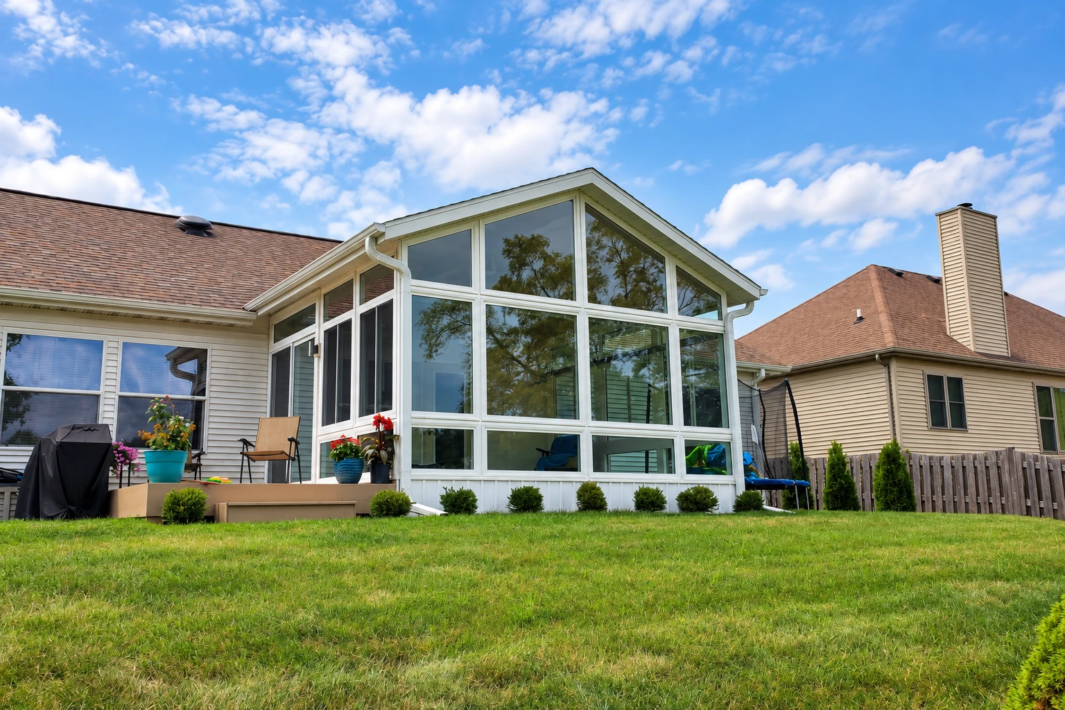 A tall, white-framed glass sunroom extends from a suburban home's deck, facing a lawn between neighboring houses.