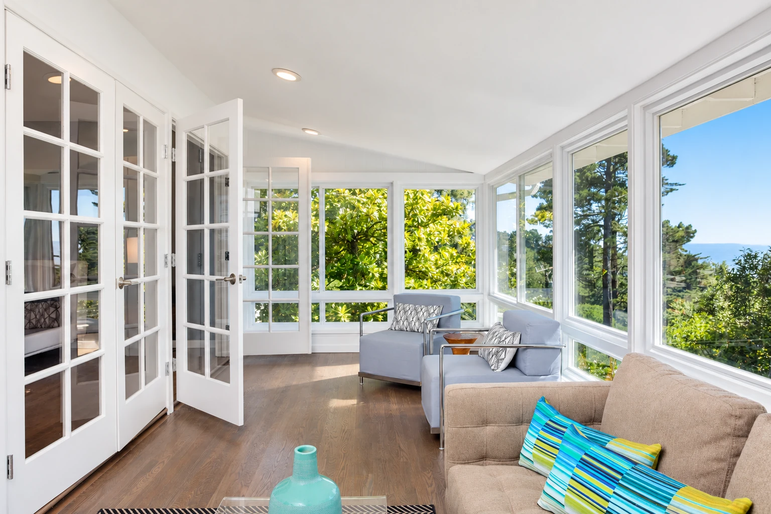 Bright four-season sunroom with expansive windows, French doors, wood flooring and neutral seating.