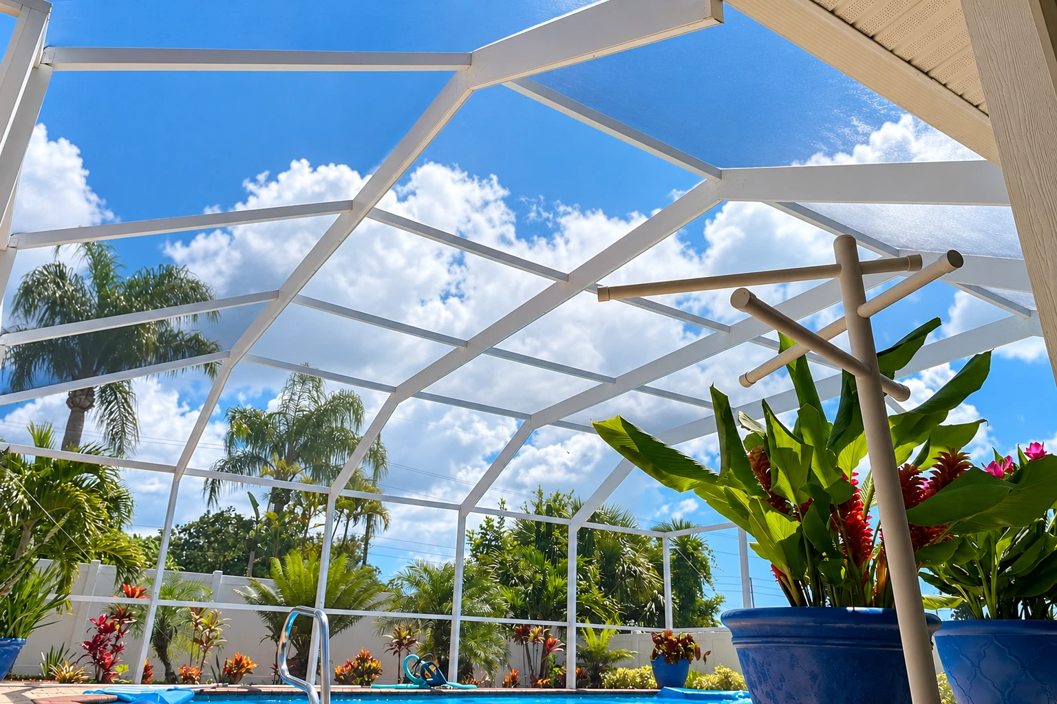 White framing and clear panels form a screened patio enclosure over a swimming pool, with tropical plants beneath a blue sky.
