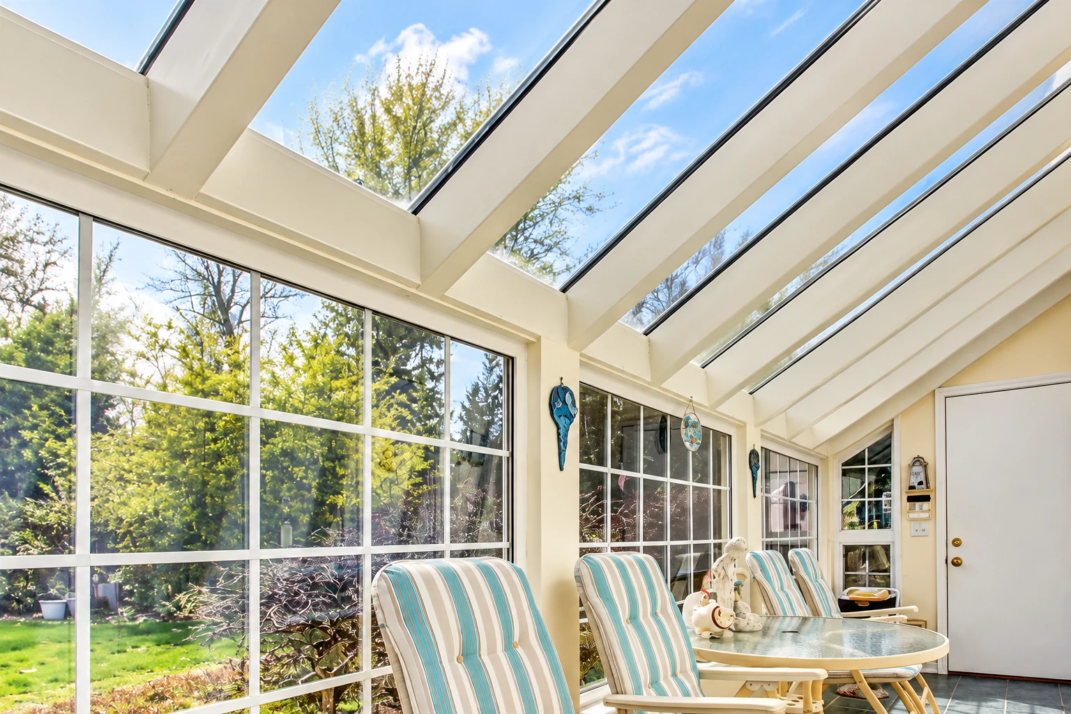 Solarium with a glass-paneled sloped roof, large windows, and striped lounge chairs overlooking a leafy yard.