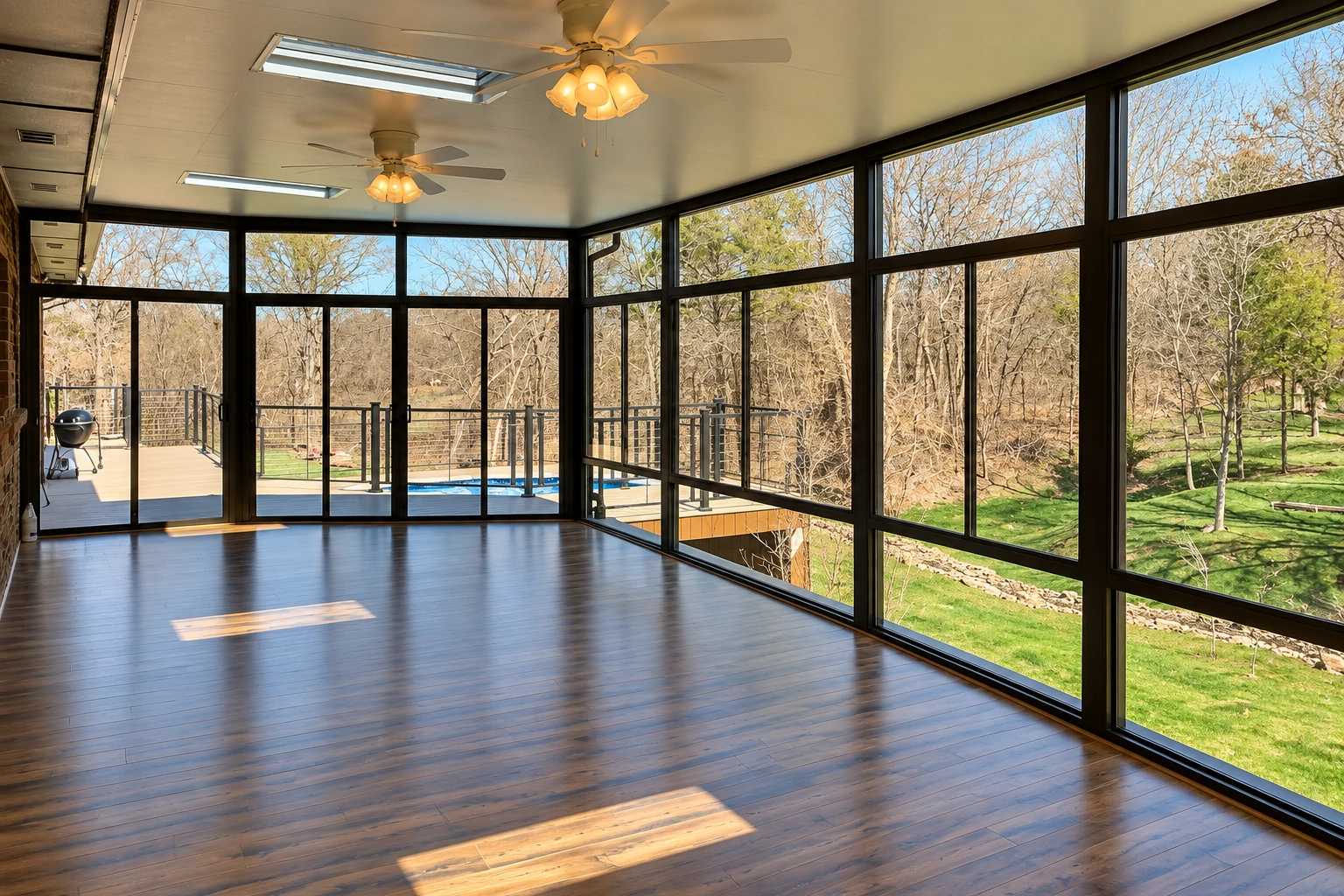 Empty sunroom with dark wood floors, black-framed windows, and views of a wooded yard and pool.