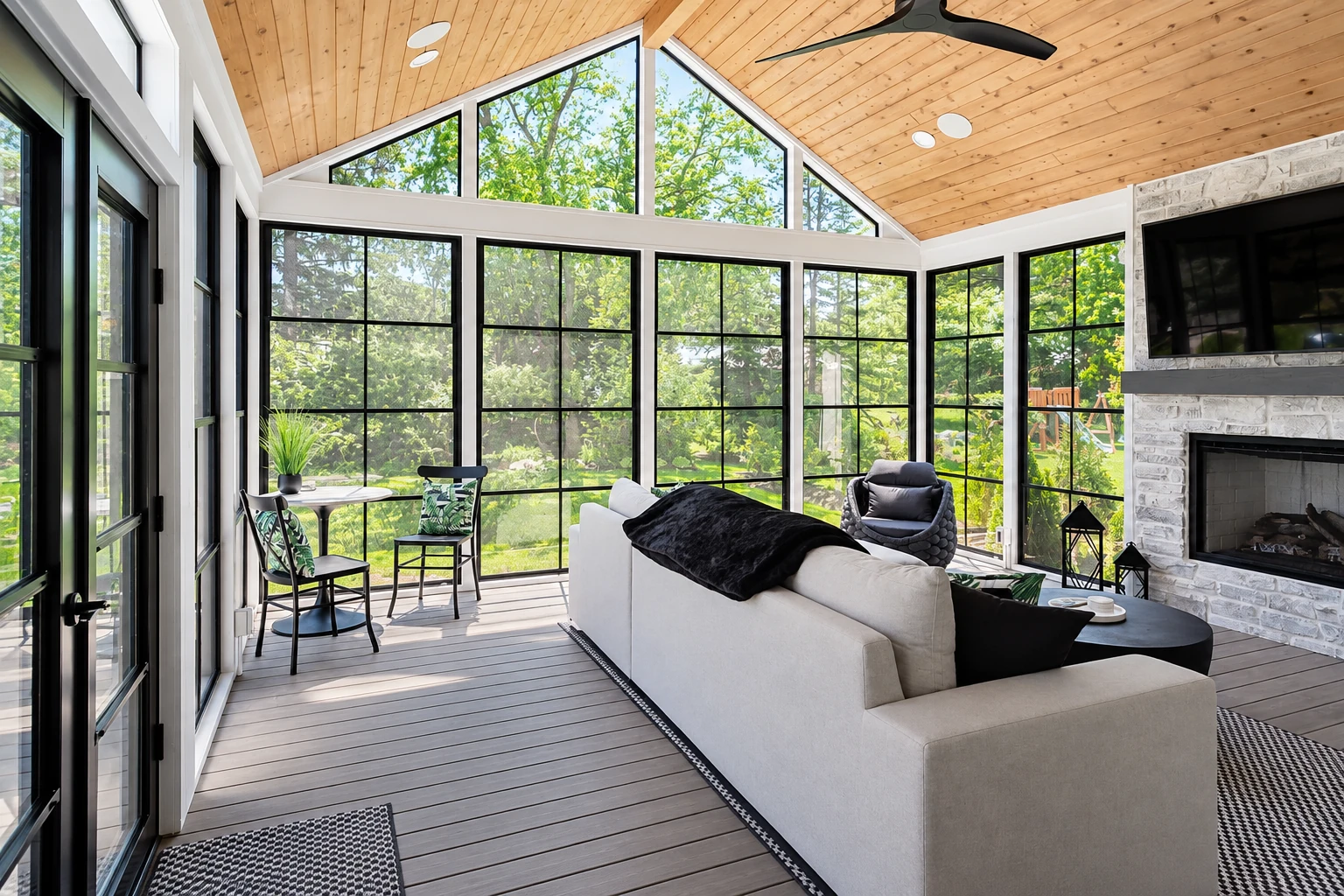 Three-season sunroom with a wood-plank ceiling, black-framed windows, a stone fireplace and a seating area.